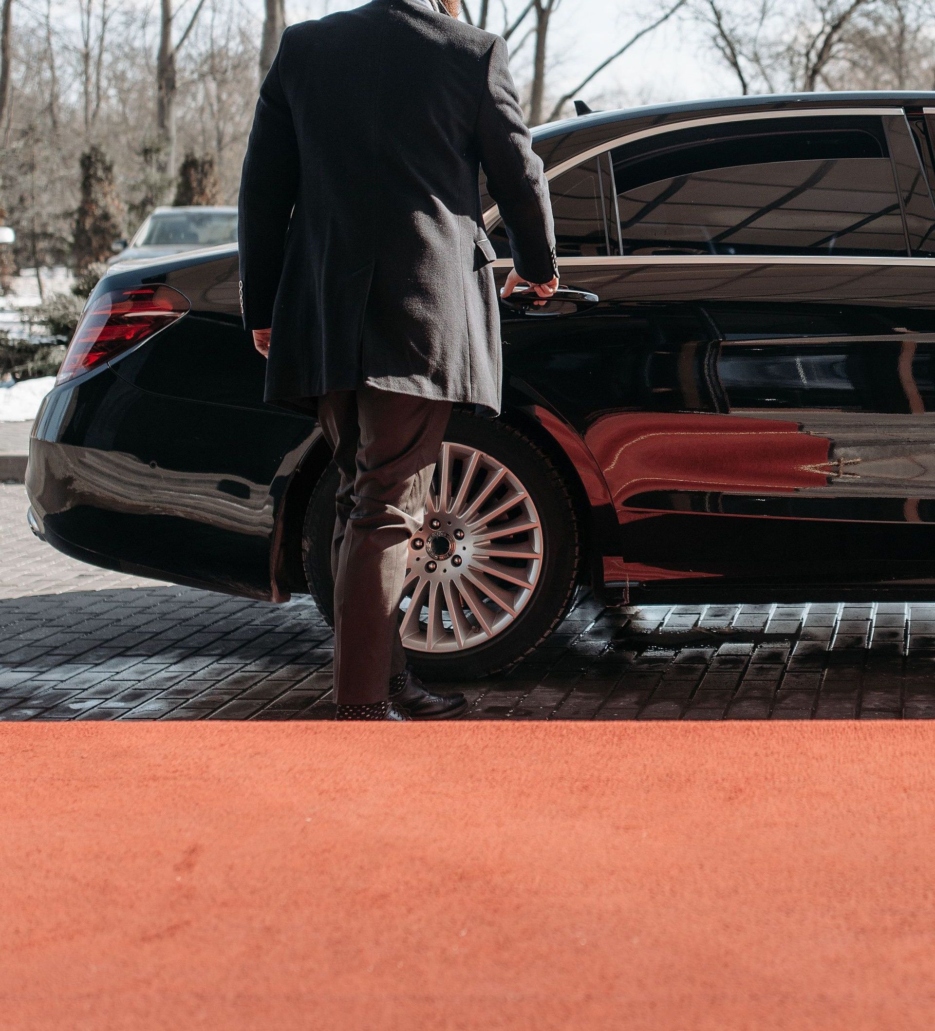 Bald man in a black suit approaches a luxury black car on a sunny day.